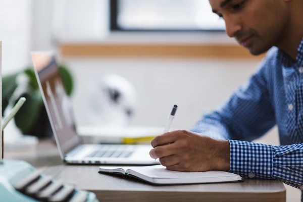 Stock image of a man in a blue-checked shirt writing in a notebook next to a laptop. 