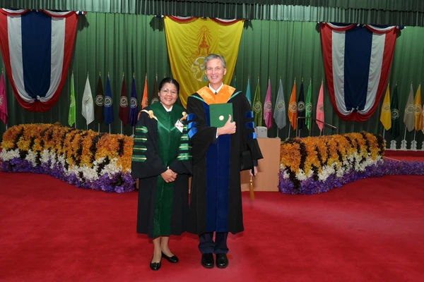 Brad Cardinal stands next to Thai colleague Amphorn Sriyapai in front of colorful decorations and holding his honorary doctorate.