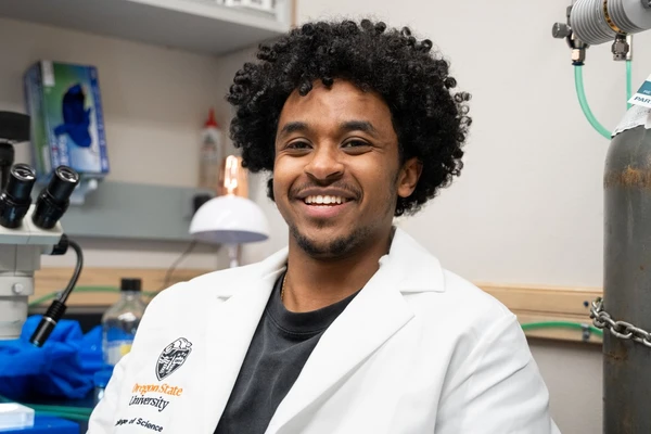 Estifanos Berhe wearing a white lab coat sitting in a university lab and smiling.