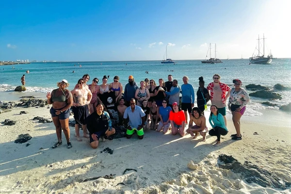 A group of students in swimsuits and sun-protective clothing pose with instructor Itchung Cheung on a sandy beach in front of beautiful turquoise Caribbean water.