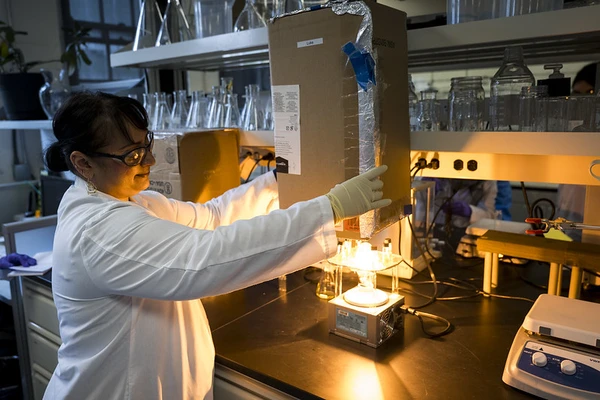 Marilyn Rampersad Mackiewicz wearing a lab coat and goggles as she works on something very very bright on a lab bench.