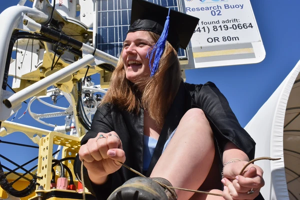 Athena Abrahamsen sitting on a research buoy in her graduation cap and gown. Photo credit Luke Hunter.