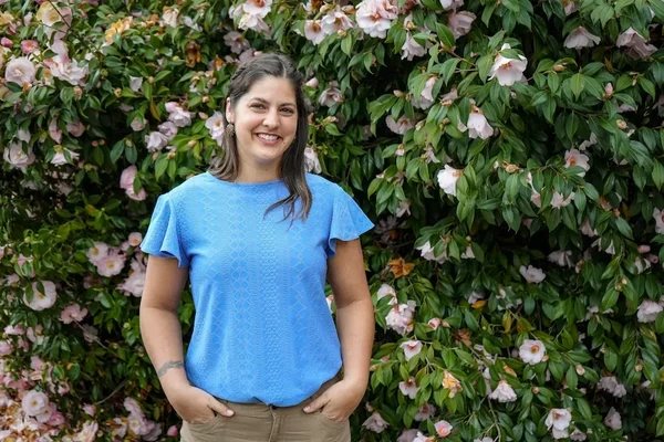 Rebecca Wheaton, smiling brunette woman, in a blue t-shirt in front of a flowering shrub.
