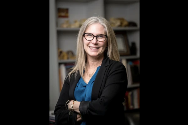 Distinguished Professor Melissa Cheyney stands in her office wearing black cardigan and blue blouse, smiling at the camera.