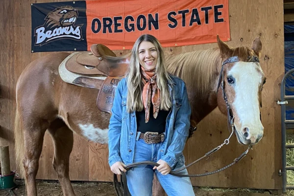 Abigail Roth stands in front of a brown and white horse and an OSU banner, wearing denim and smiling.