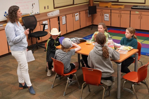 A teacher addresses five kids seated around a table in a classroom.