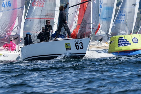 A small team of young people dressed in black sail their white boat at the front of a group of colorful sailboats on dark blue water.