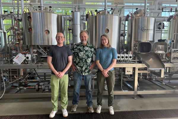 A young man, an older man, and a young woman smile as they stand in front of the large metal vats in OSU's pilot brewery.