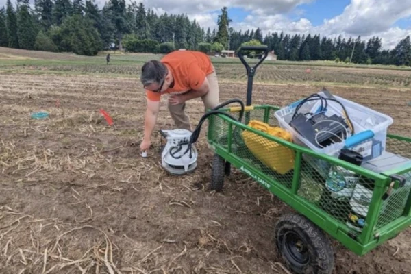 Christian Lessey wears an orange shirt while stooping to test something in a field.