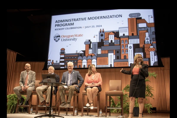 OSU administrators sit in chairs onstage at PRAx with a PowerPoint presentation on screen behind them announcing the Administrative Modernization Program kickoff in July 2024.