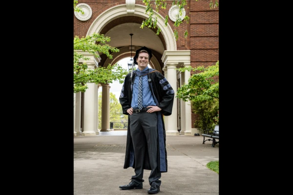 Patrick Callagy stands in front of the archway of Weatherford Hall, hands on his hips wearing his graduation regalia.