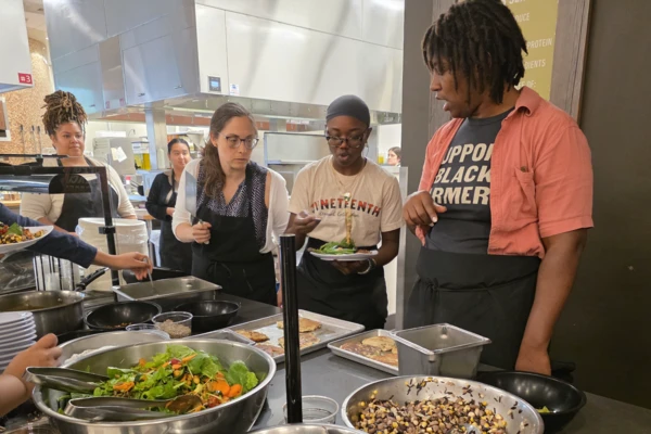 Several people gathered around a counter laden with metal bowls of ingredients inside an industrial kitchen.