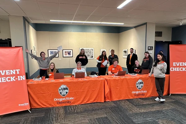 Excited students sit and stand near orange-draped tables ready to welcome visitors for an event check-in.