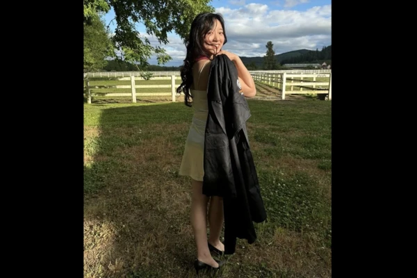 Kaitlyn Kim stands in a grassy yard looking back at the camera with her graduation gown slung over her shoulder.