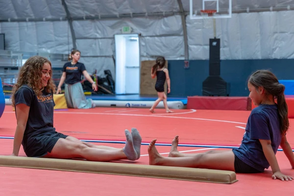 Two young women sit facing each other on a gymnastics mat with their legs stretched out in front of them.