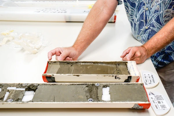 A fair-skinned man's hands gesturing to two metal trays of grayish-brown sediment.