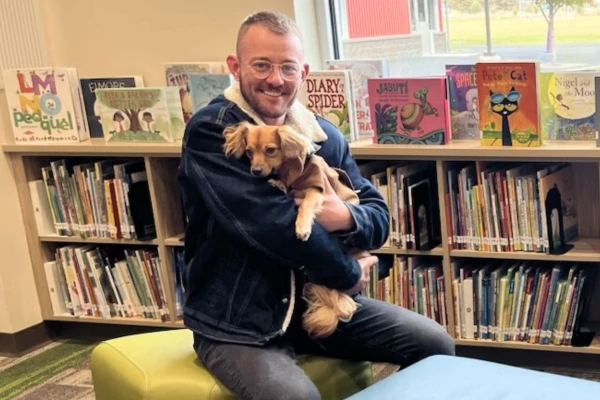 A man in a dark jacket holding a cute puppy and smiling while sitting in a children's library.