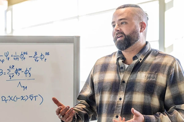 Jackson Harter in a plaid shirt in front of a whiteboard covered in mathematical formulas.