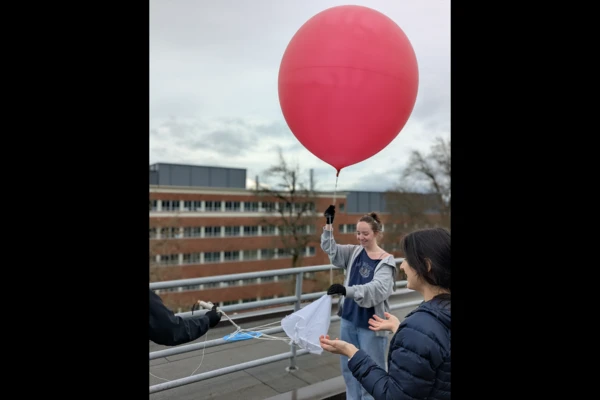 A student holds onto the tail of a big red weather balloon on the roof of an OSU building pre-launch.