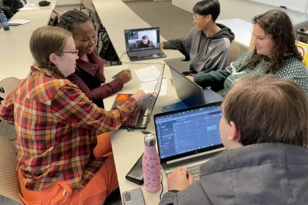 A group of business students sit around a narrow table with their laptops working together.