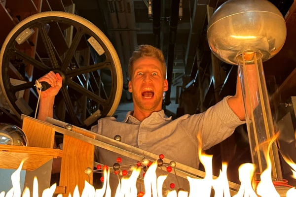 A mad scientist holds some physics instruments aloft behind a row of small flames, looking very dramatic.
