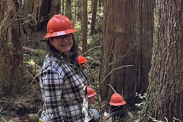 Woman in orange hard hat and flannel shirt smiling in the forest.