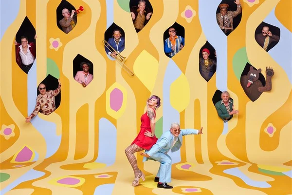 A creative photo of the band Pink Martini, with the lead pianist and singer standing on a psychedelic yellow painting in the foreground and various band members popping out of windows above them.